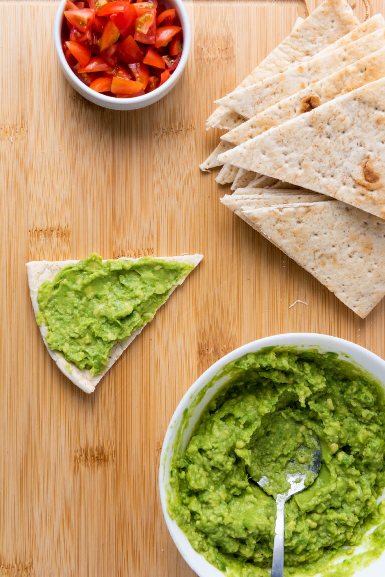 mashed avocado spread on pita triangles, tomatoes in a bowl on a wooden table