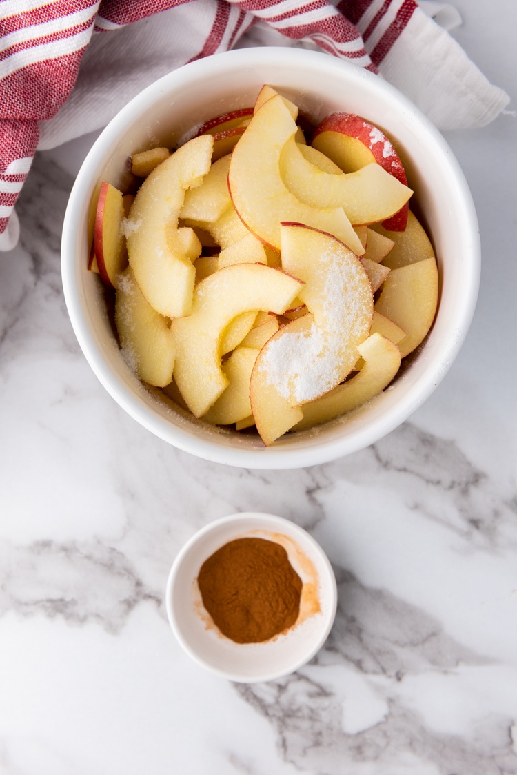 A picture of sliced apples in a bowl next to a bowl of cinnamon.