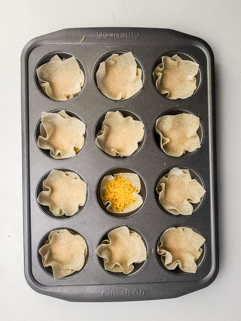 A picture of egg roll wrappers stuffed into cupcake tins with shredded cheese topping one wrapper.