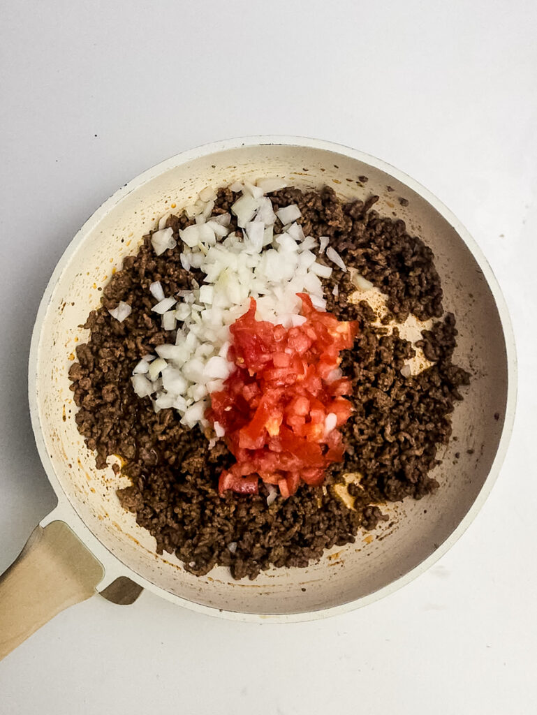A picture of cooked ground beef in a skillet with chopped onion and diced tomatoes.