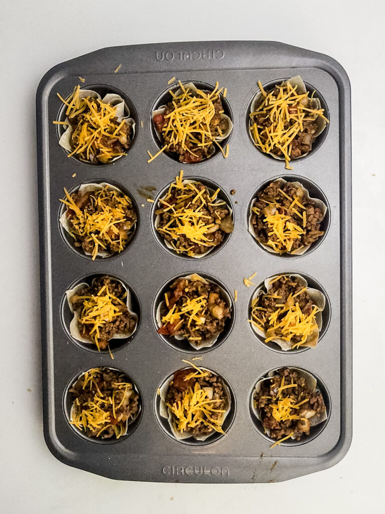 A picture of egg roll wrappers filled with a ground beef mixture topped with shredded cheese in cupcake tins.