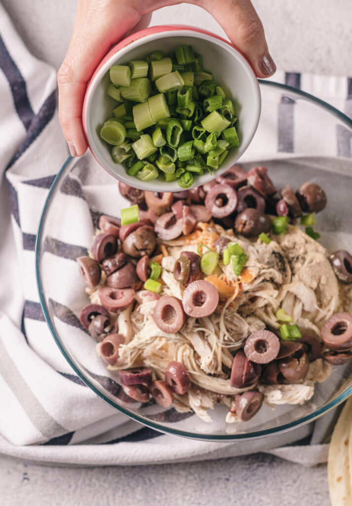 A picture of green onions being poured into a bowl with a chicken, cream cheese, olive mixture.