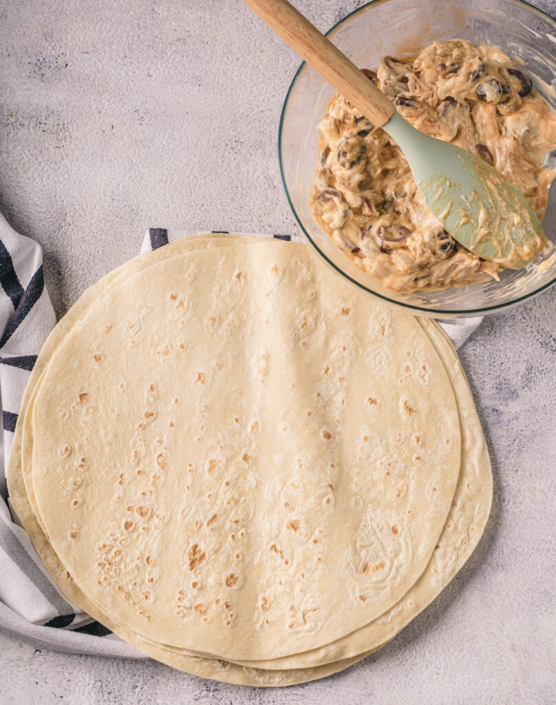 A picture of several tortillas stacked on top of one another near a bowl holding a chicken and cream cheese mixture and a spatula.