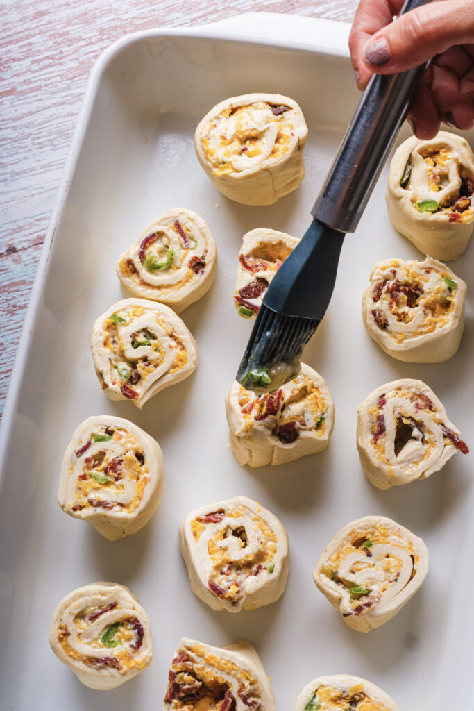 A picture of garlic butter being brushed over uncooked jalapeño pinwheels in a baking pan.