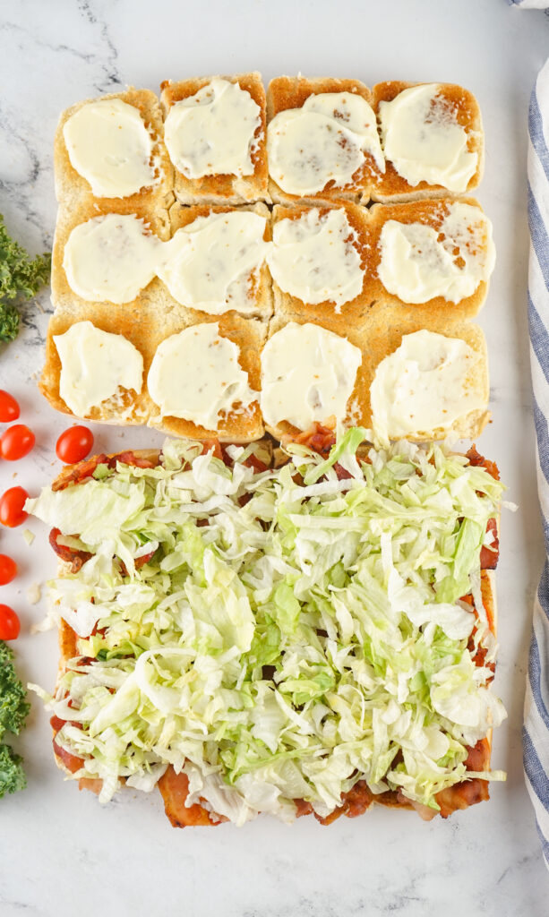 A picture of toasted sliced slider buns, topped with mayonnaise, tomato, bacon, and lettuce.