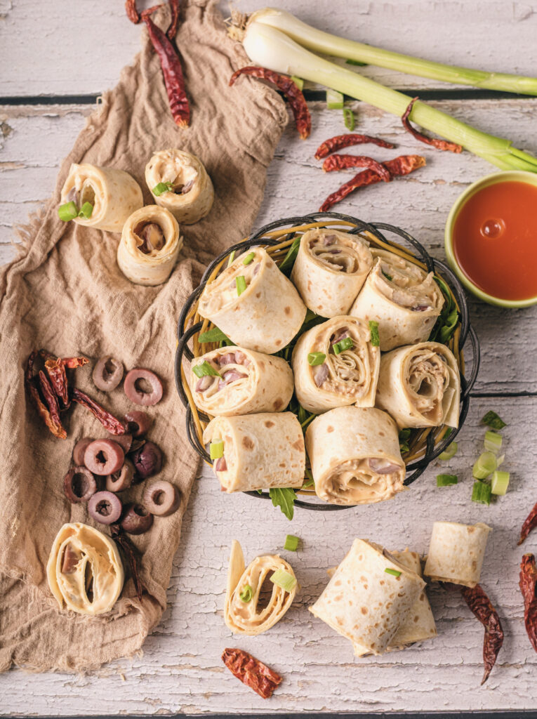 A picture of buffalo chicken pinwheel sandwiches stacked together in a bowl, surrounded by more sandwiches and ingredients.