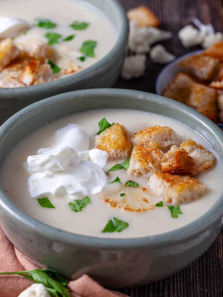 A picture of cauliflower soup in a bowl with sour cream, croutons, and parsley.