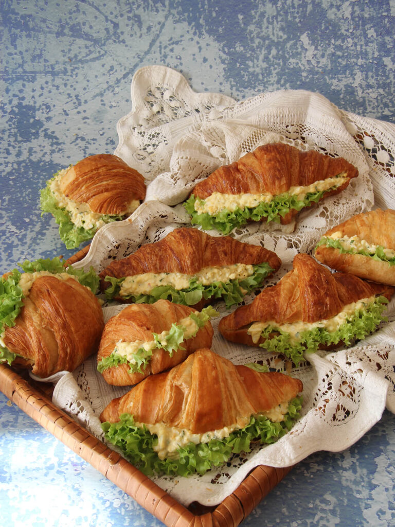 A picture of a collection of croissant egg salad sandwiches in a basket with white cloth.