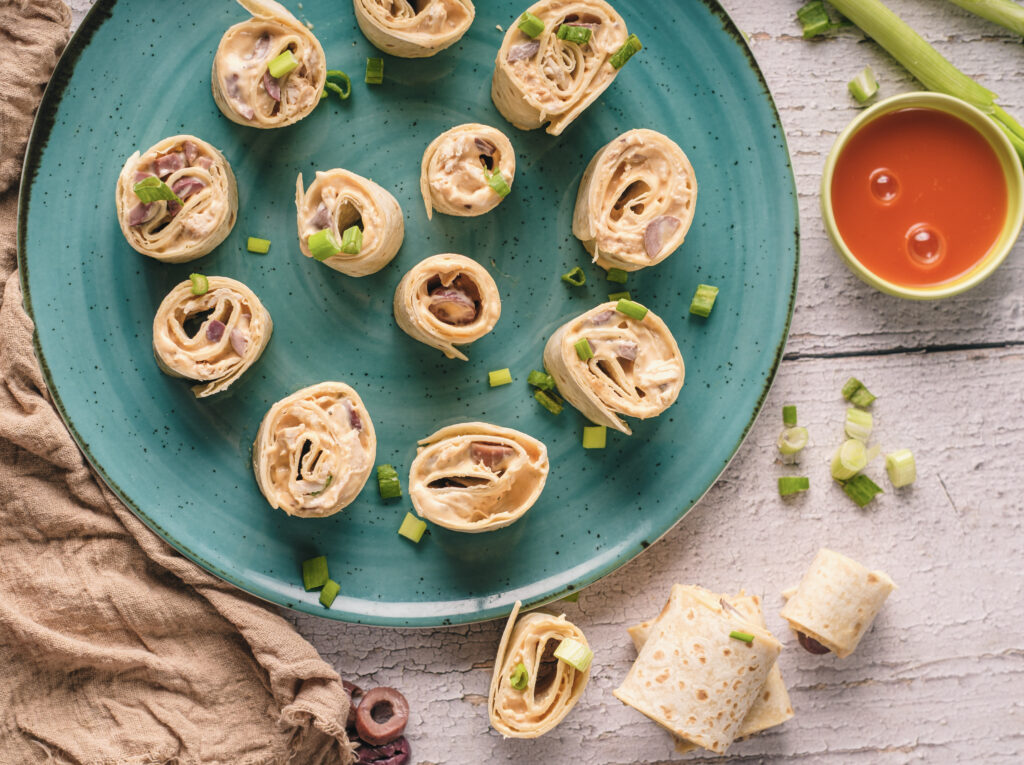 A picture of buffalo chicken pinwheel sandwiches on a plate with buffalo sauce on the side.