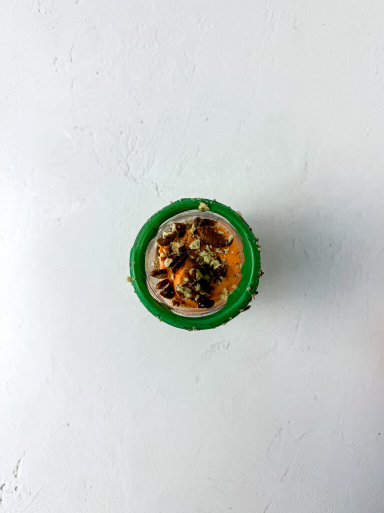 An overhead shot of a carrot cake muffin topped with orange frosting and pecans in a jar.