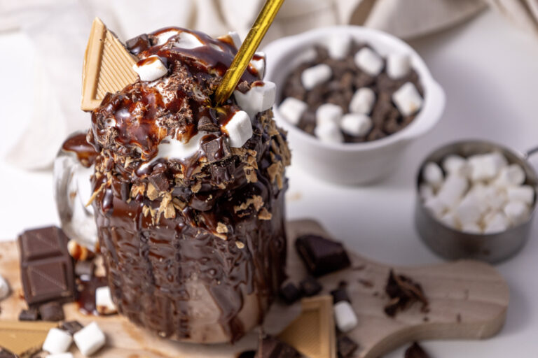 A close up picture of a mug of chocolate shake topped with pieces of chocolate and marshmallows.