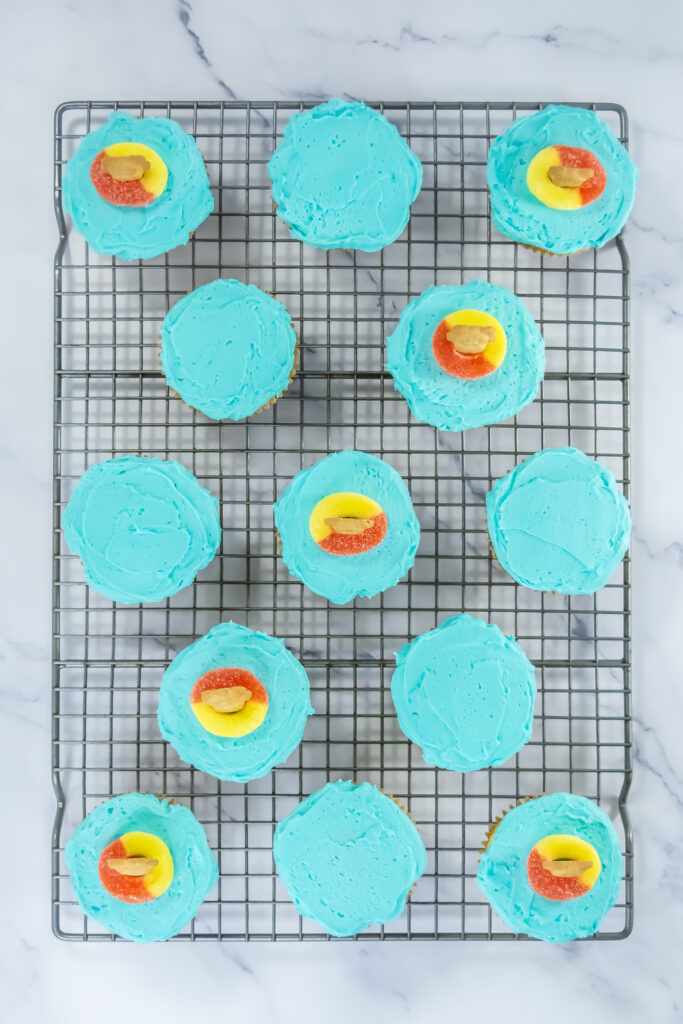 A picture of thirteen blue frosted cupcakes, with seven of them topped by a Teddy Graham cookie placed in the middle of a peach ring candy.