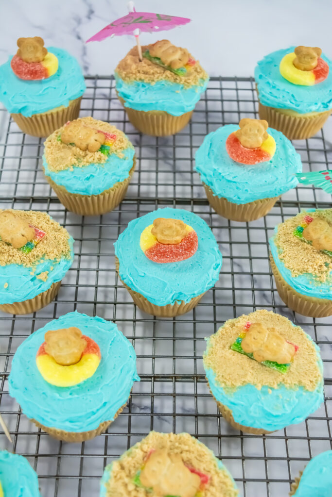 A picture of blue frosted cupcakes topped with candy and Teddy Graham cookies, garnished with a mini umbrella.