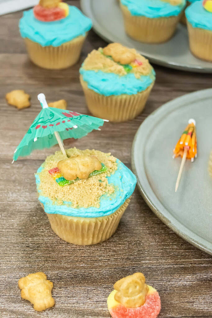 A picture of a blue frosted cupcake decorated by a Teddy Graham cookie on a cookie crumb beach with a mini umbrella.