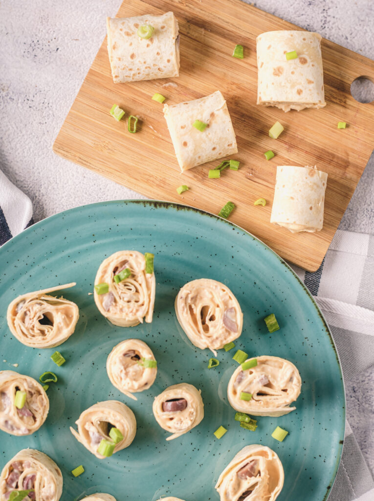 A picture of buffalo chicken pinwheel sandwiches on a plate and on a cutting board, garnished with sliced green onions.