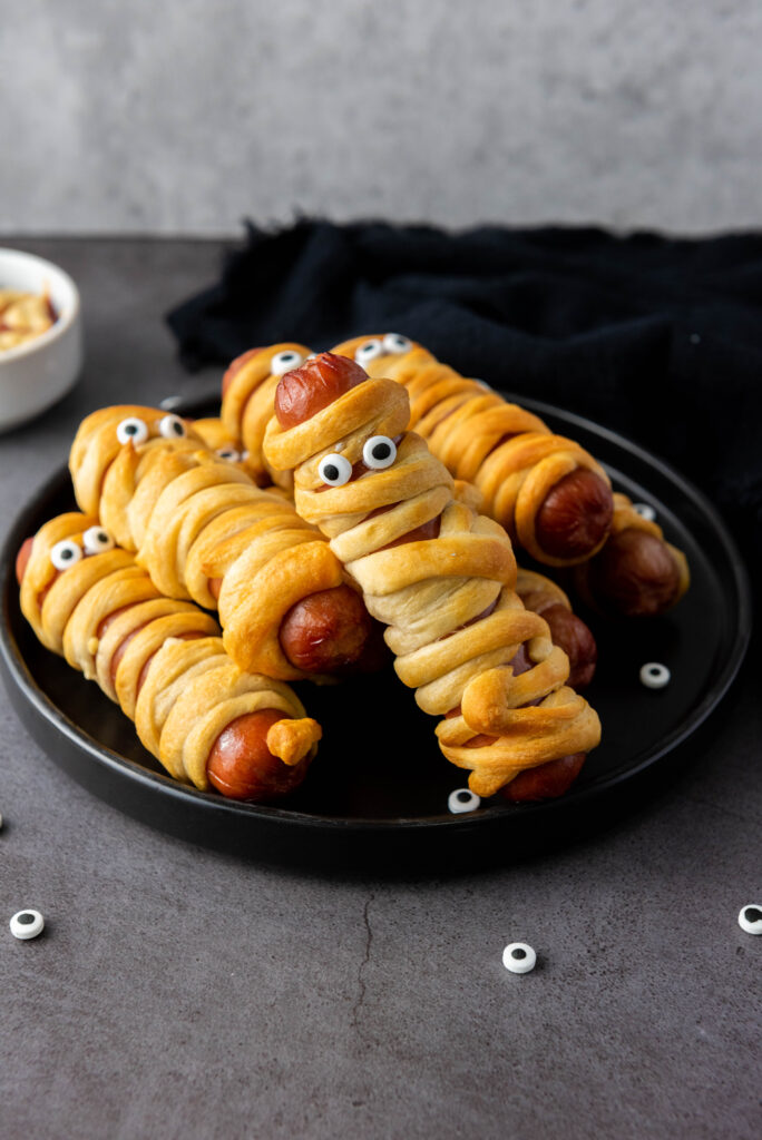 A picture of a pile of finished Halloween mummy dogs on a black plate.