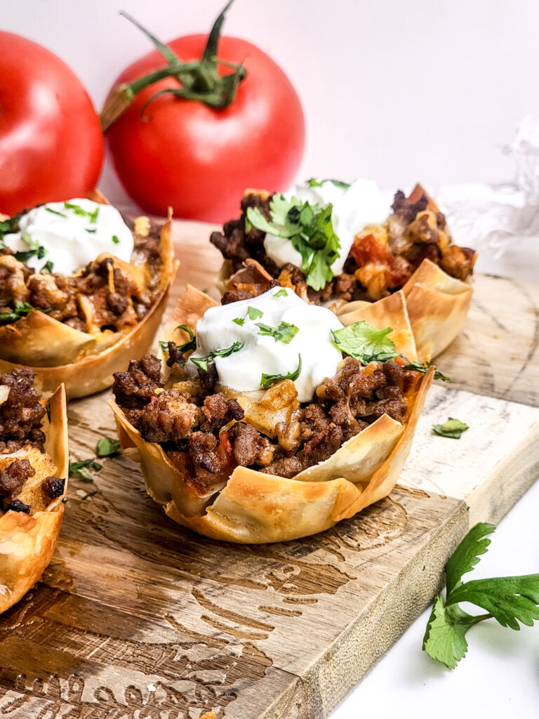 A picture of mini taco cups topped with sour cream and cilantro displayed on a wooden cutting board.