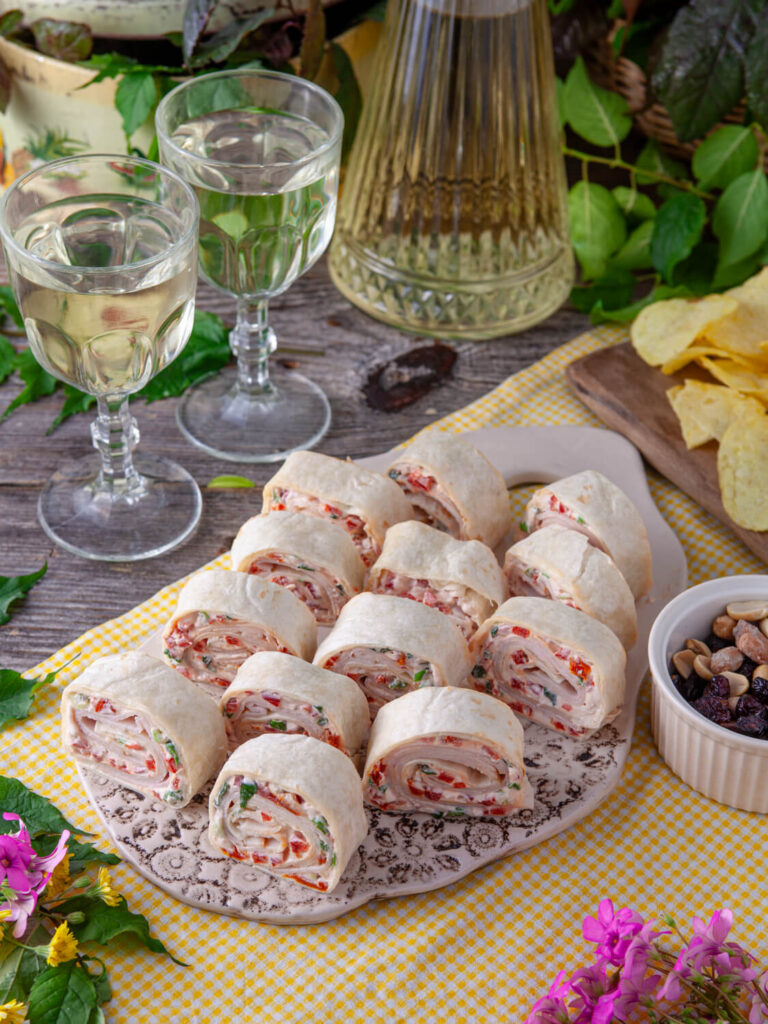A picture of three rows of individually cut turkey tomato pinwheel sandwiches on a platter with wine in the background.