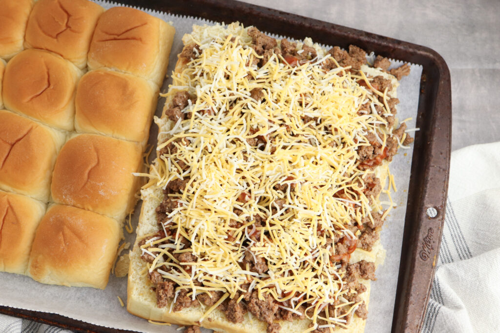 A picture of ground beef and shredded cheese spread over Hawaiian rolls which have been cut in half.