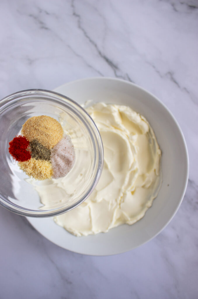 A picture of spices being poured into a bowl of cream cheese.