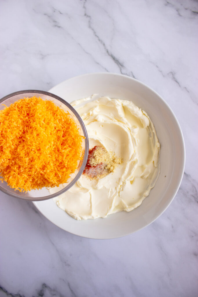 A picture of shredded cheddar cheese being poured into a bowl with whipped cream cheese and spices.