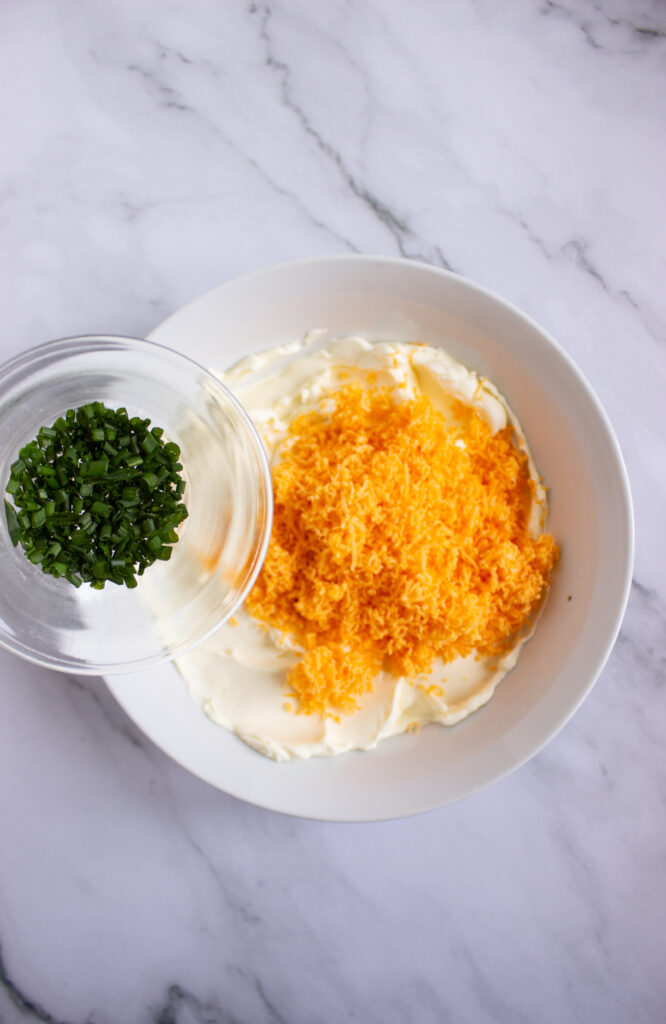 A picture of chives being poured into a bowl with whipped cream cheese, shredded cheddar cheese, and spices.