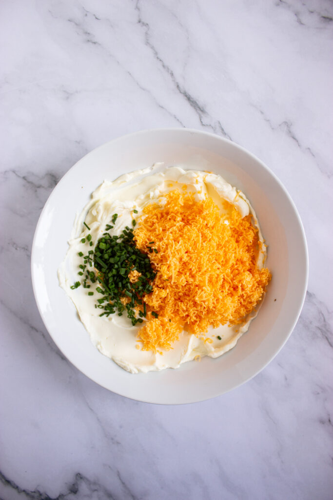 A picture of chives, whipped cream cheese, shredded cheddar cheese, and spices together in a bowl, unmixed.
