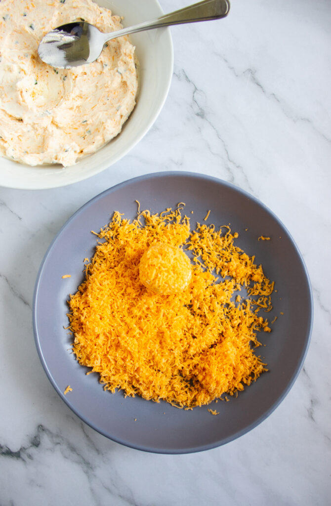 A picture of a sesasoned cream cheese ball being rolled in shredded cheddar cheese.