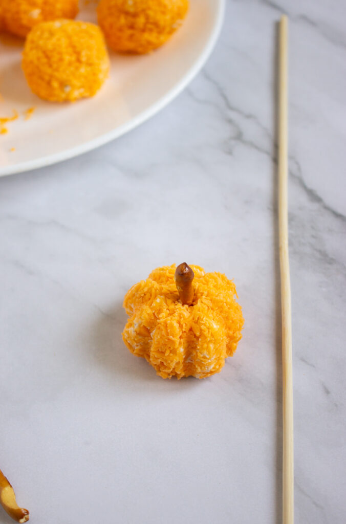 A picture of a cheese ball shaped like a pumpkin, rolled in cheddar cheese with a pretzel sticking out of the top to resemble a pumpkin.