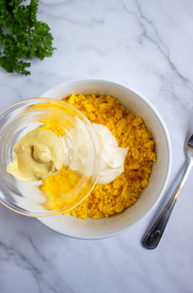 A picture of mustard about to be poured into a bowl of mayonnaise and crumbled hard boiled egg yolks.