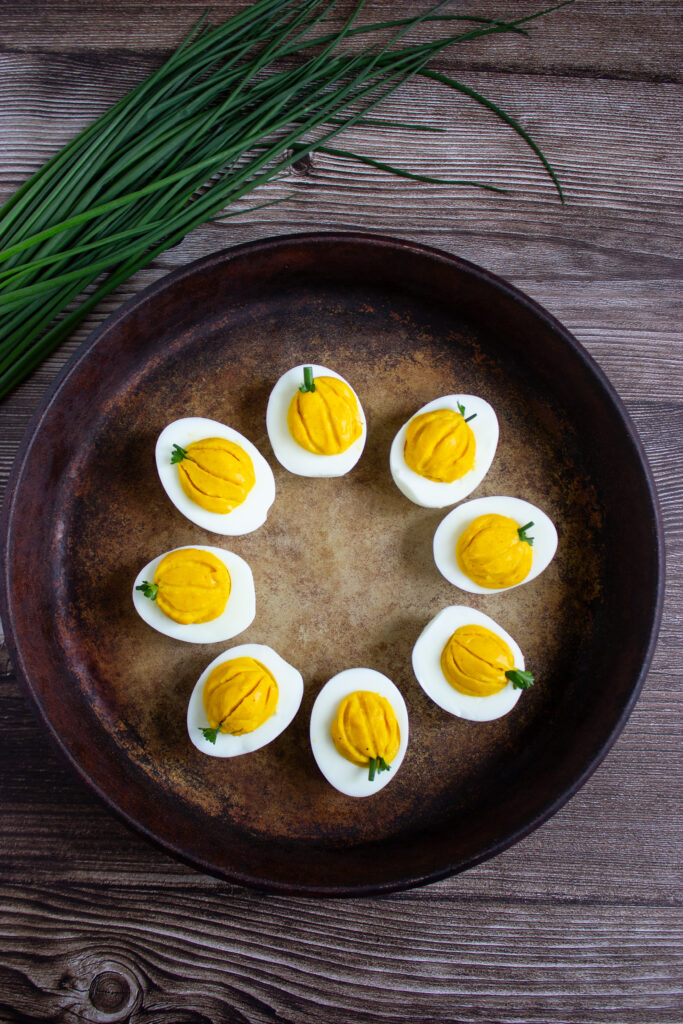 A picture of a set of deviled eggs in a pan, with grooves pressed into the yolks and a small slice of chive placed at the top to make the yolk resemble a pumpkin.