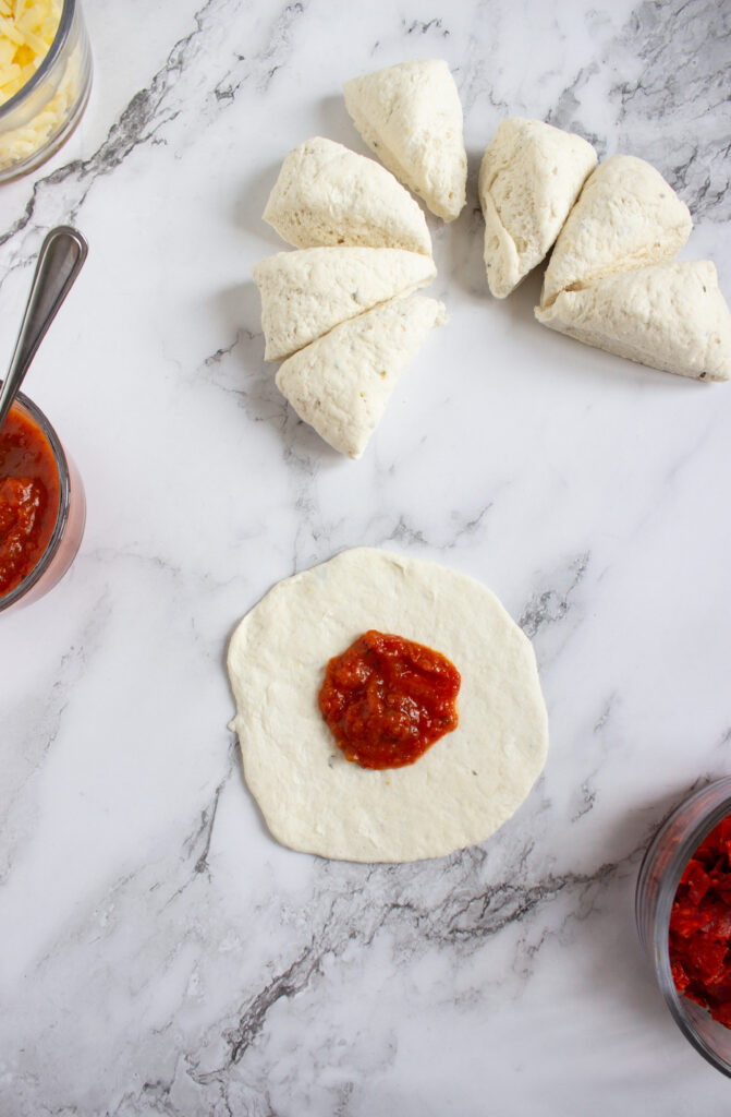 A picture of flattened bagel dough topped with pizza sauce.