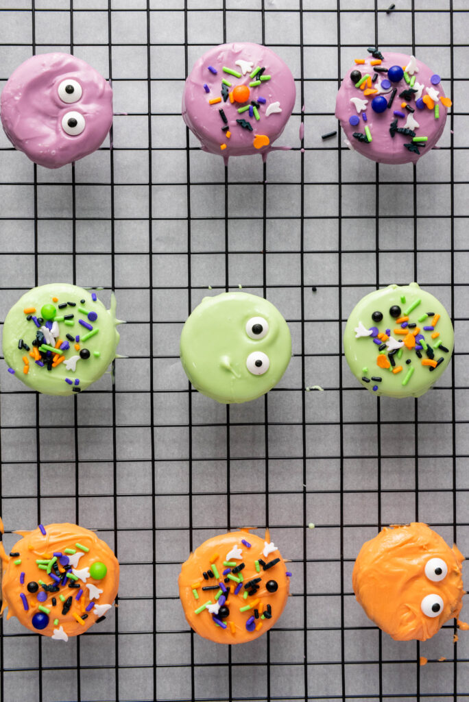 A picture of three rows of oreos dipped in purple, green, and orange melted candy, topped with Halloween decorations and sprinkles.