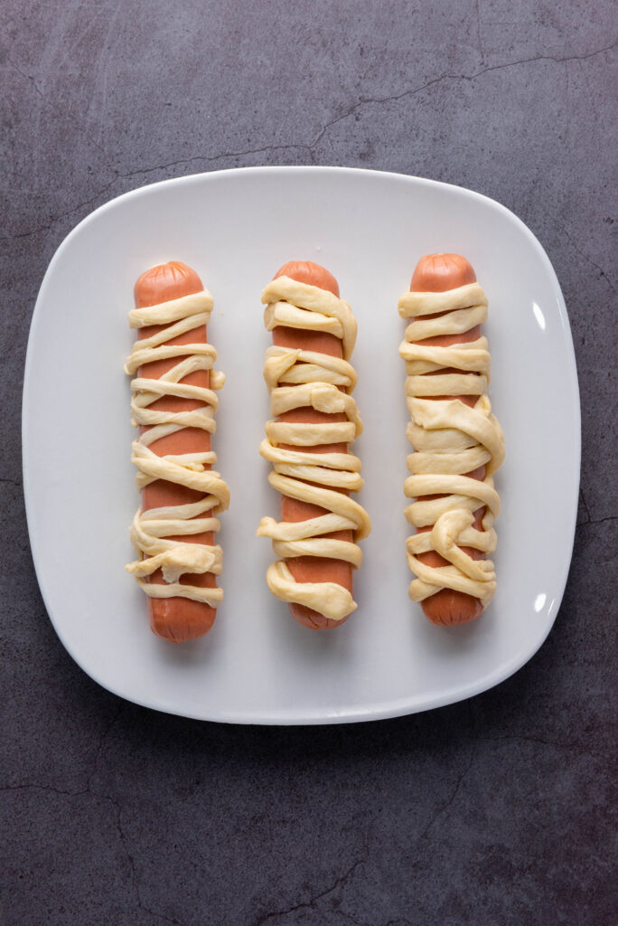 A picture of three hot dogs wrapped in crescent roll dough, uncooked.
