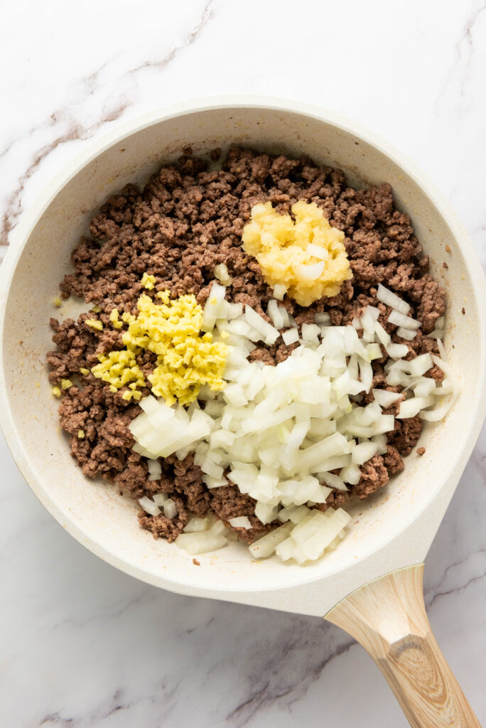 A picture of cooked ground beef in a skillet with uncooked minced garlic, diced onion, and ginger.