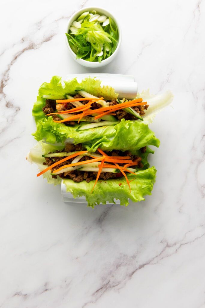 A picture of two butter lettuce leaves topped with kimchi, seasoned ground beef, sliced cucumber, and slivers of carrots.