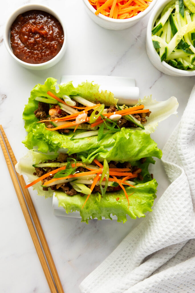 A picture of two butter lettuce leaves topped with kimchi, seasoned ground beef, sliced cucumber, slivers of carrots, chopped green onions, and sesame seeds.