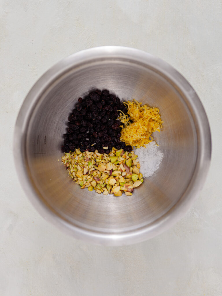 A picture of chopped pistachios, blackcurrants, lemon zest, olive oil and salt in a tin bowl, unmixed.