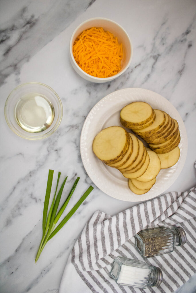 A picture of potato slices on a plate bordered by shredded cheese, olive oil, and chives on a table.