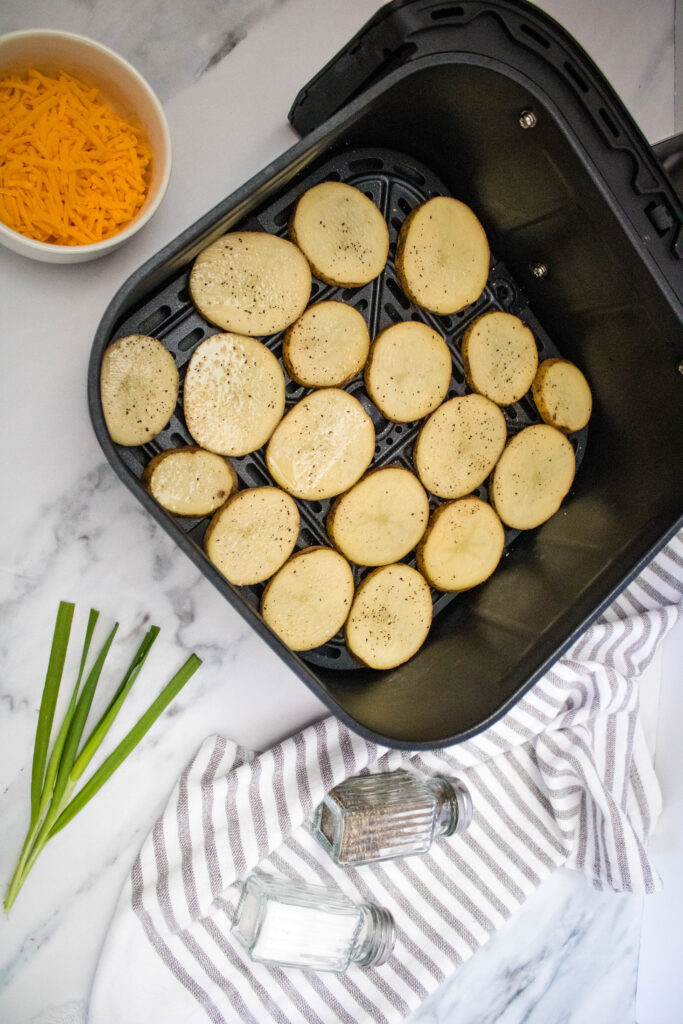 A picture of potato slices arranged in a single layer in an air fryer basket, seasoned with salt and pepper..
