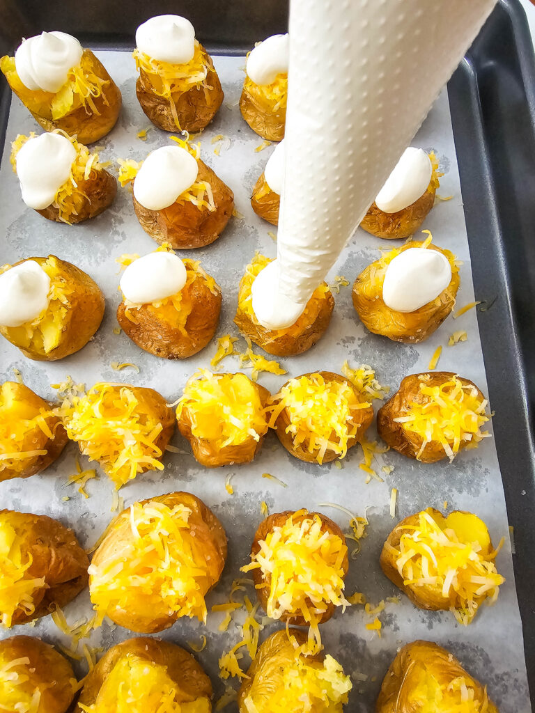 A picture of cooked mini potatoes being topped with sour cream out of a piping bag.