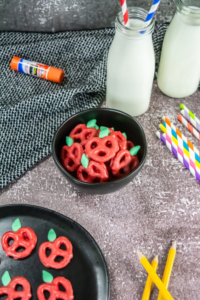 A picture of a bowl of candy coated apple pretzels surrounded by school supplies and a glass of milk.