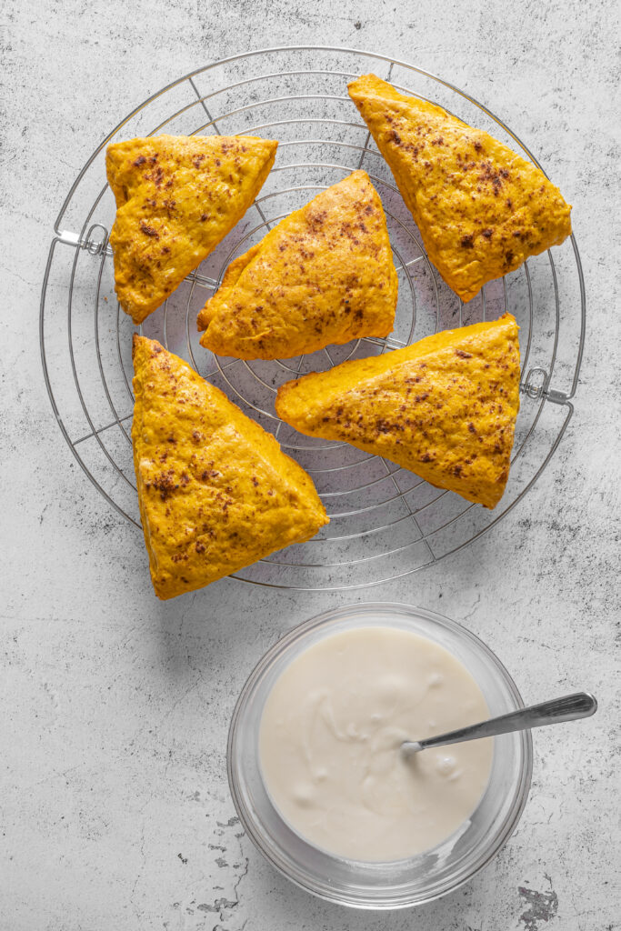 A picture of baked pumpkin pie spice scones on a wire rack next to cream glaze in a glass bowl.