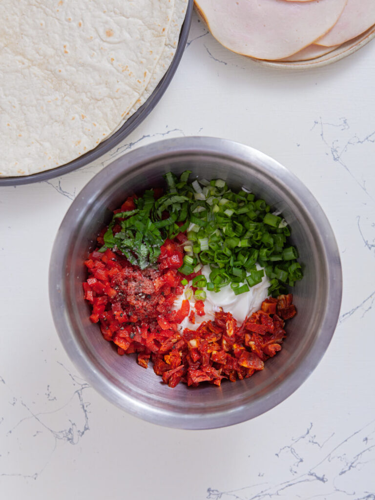 A picture of cream cheese, sour cream, sun-dried tomatoes, bell pepper, scallions, and basil unmixed n a bowl.