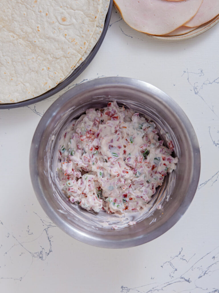 A picture of cream cheese, sour cream, sun-dried tomatoes, bell pepper, scallions, and basil mixed together in a bowl.