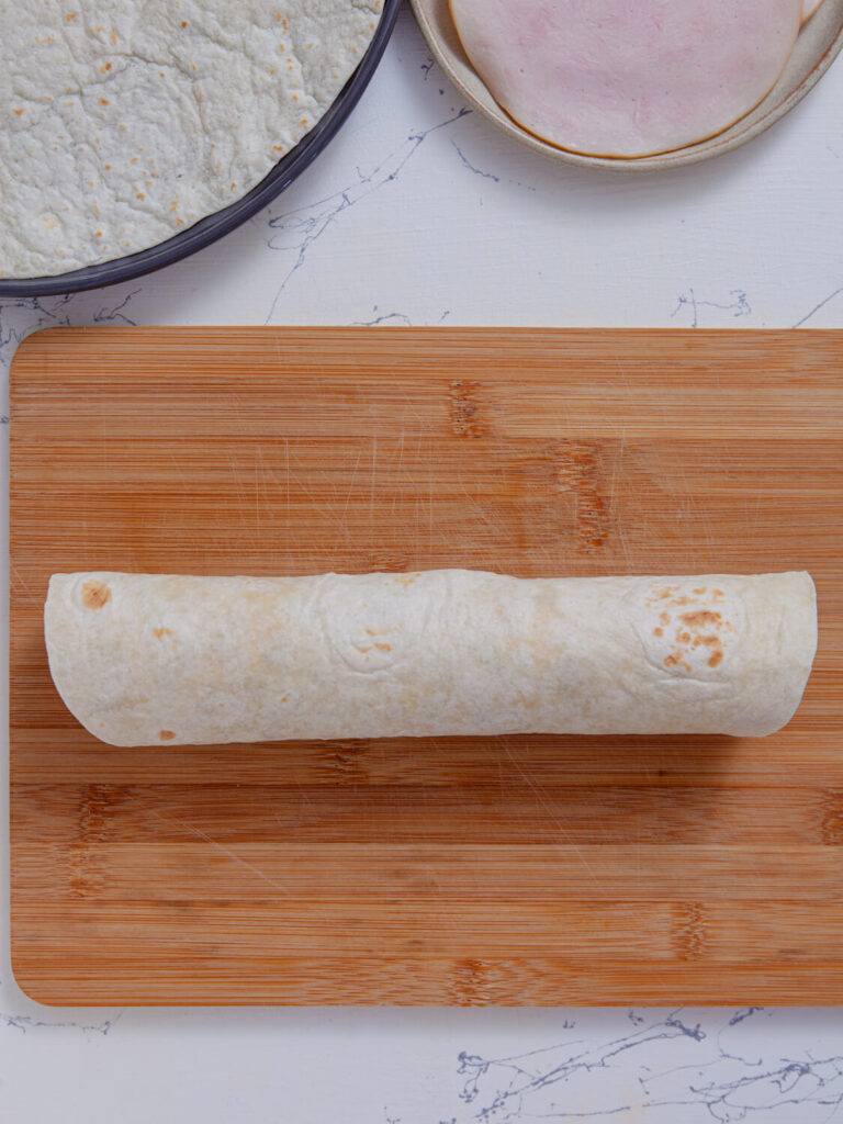A picture of a tortilla topped with a turkey tomato cream cheese mixture fully rolled into a log.