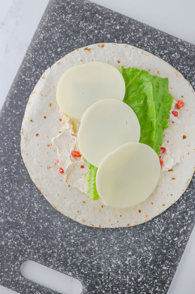 A picture of a flour tortilla topped with a cream cheese mixture, romaine lettuce leaf, and three slices of provolone cheese..