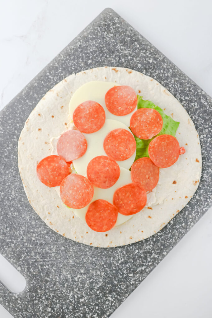 A picture of a flour tortilla topped with a cream cheese mixture, romaine lettuce leaf, provolone cheese, and twelve slices of pepperoni.