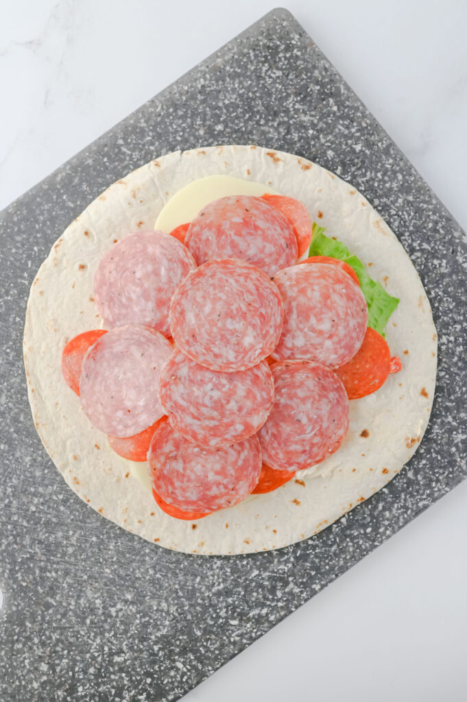 A picture of a flour tortilla topped with a cream cheese mixture, romaine lettuce leaf, provolone cheese, slices of pepperoni, and slices of salami.